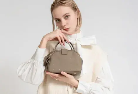 白の大きなリボンタイブラウスを着た女性モデルが、スタッズ付きのグレージュのミニバッグを両手で抱えている。